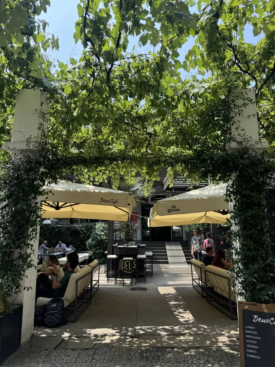 Entrance to deus cafe with sun shinning trees everywhere and two yellow umbrella's with the deus logo. People sitting down enjoying their food.