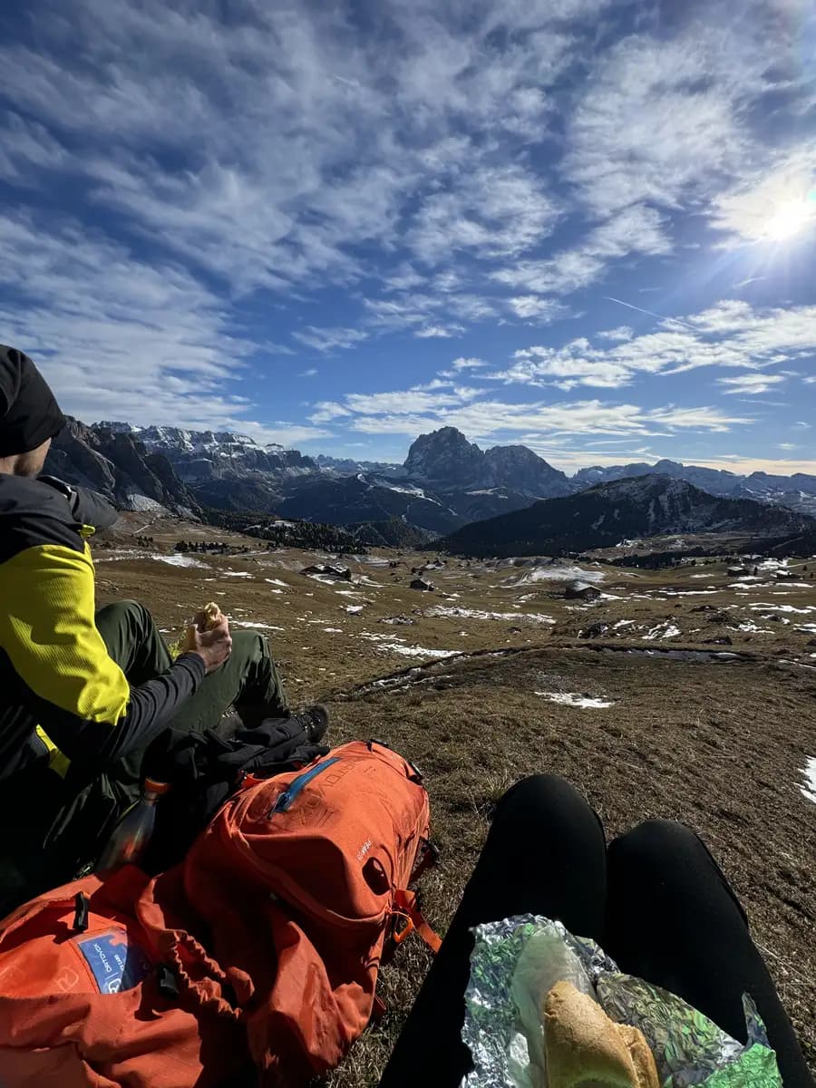Two hikers sitting on a flat rock and eating paninis while looking out over the Seceda valley in the Italian Dolomites. The background shows the massive peaks of the Sassolungo and the Sella group mountain ranges under a clear sky.