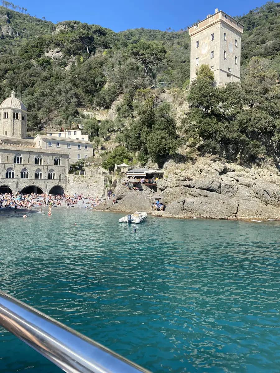 The historic medieval San Fruttuoso Abbey sitting directly on a small pebbly beach in a secluded cove in Liguria, Italy, surrounded by clear turquoise water.