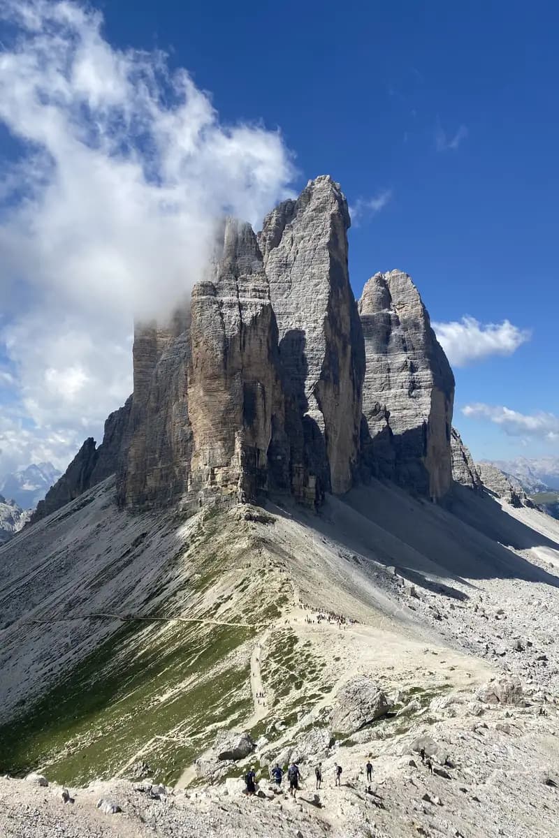 A clear view of the Tre Cime di Lavaredo peaks from the Forcella Lavaredo saddle in the Italian Dolomites. The three massive limestone towers rise against a blue sky above the rocky alpine hiking trail.