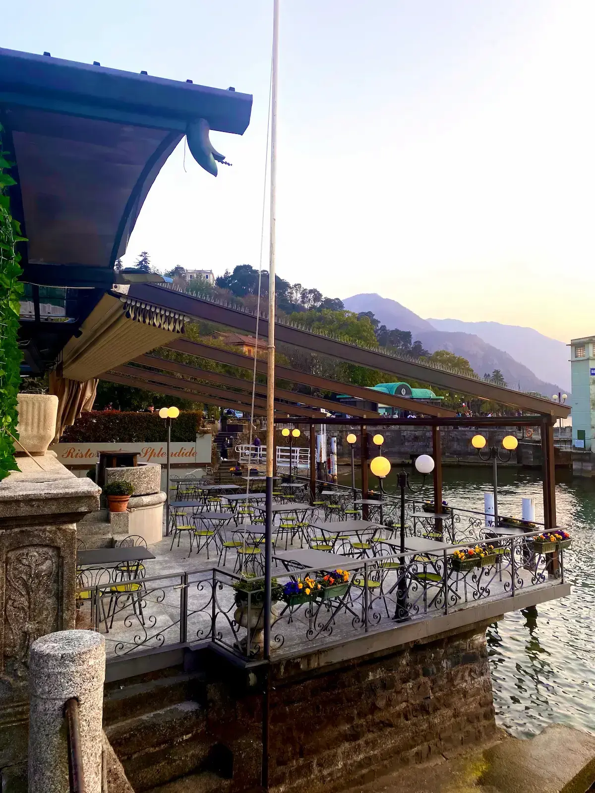 Waterside restaurant terrace in Bellagio, Lake Como, featuring outdoor dining tables and chairs under a green canopy with a scenic view of the lake and mountains.