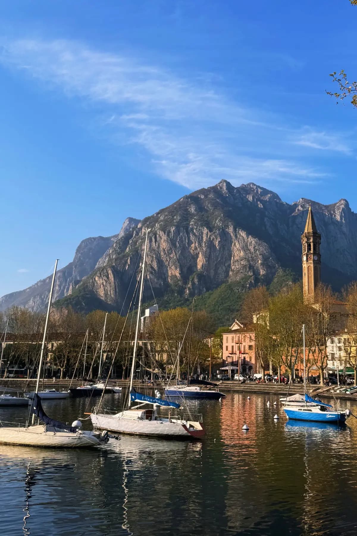 View of the Lecco waterfront at Lake Como with traditional lake boats docked near the historic tower under a clear sky.