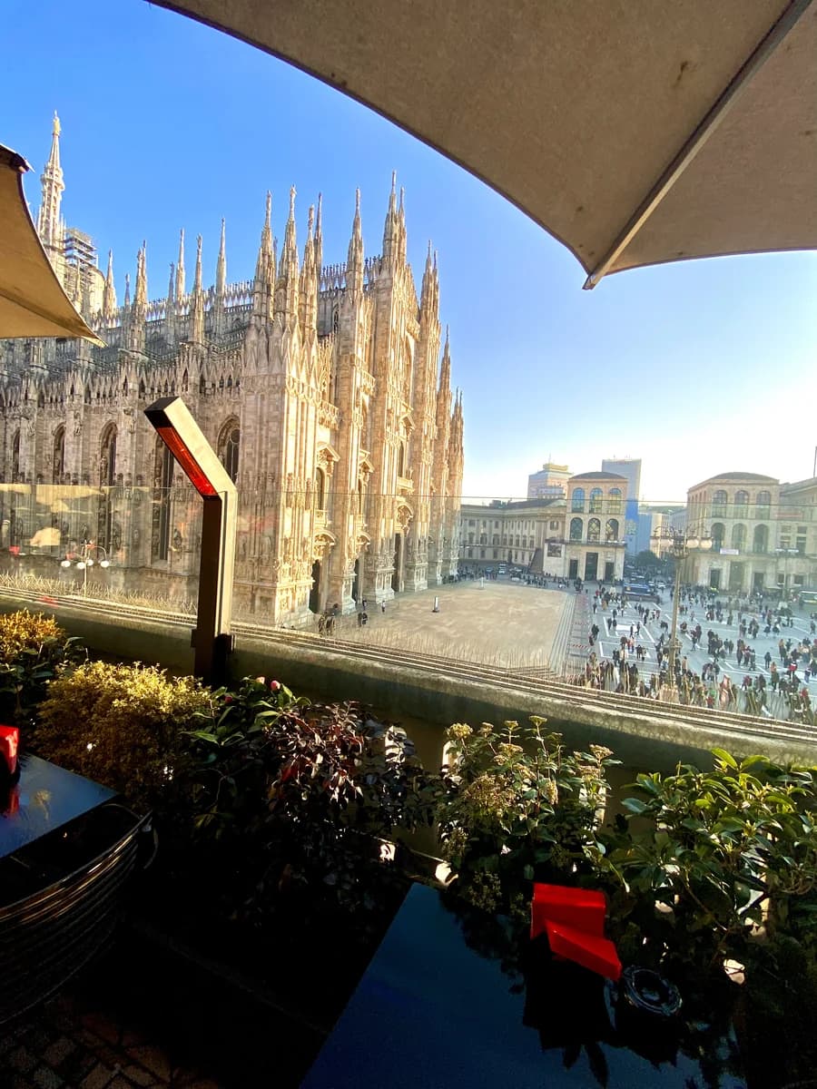 Close-up view of the intricate white marble spires of the Milan Duomo from the vibrant orange terrace of the Terrazza Aperol rooftop bar.