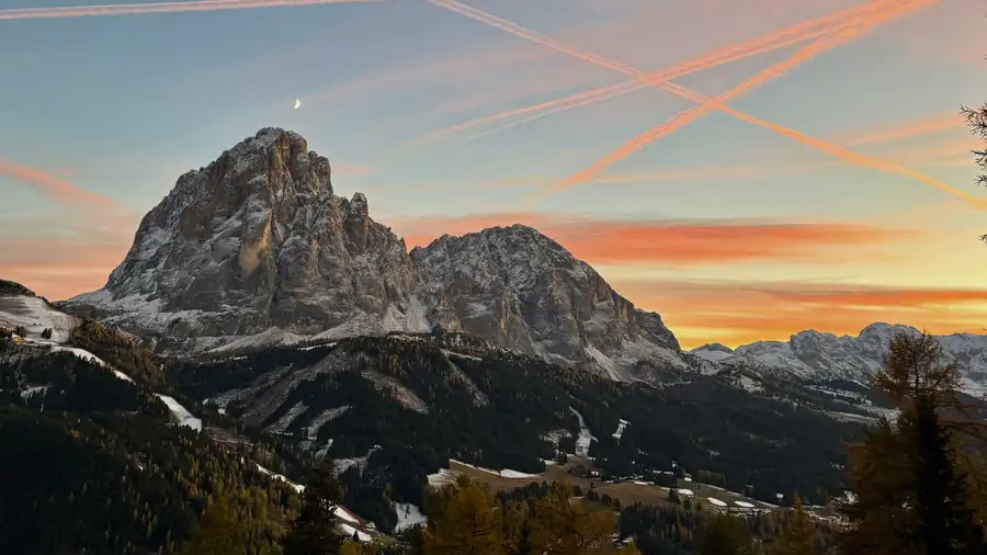 A dramatic sunset with warm golden and orange light glowing behind the jagged peaks of the Sassolungo mountain group in the Italian Dolomites.
