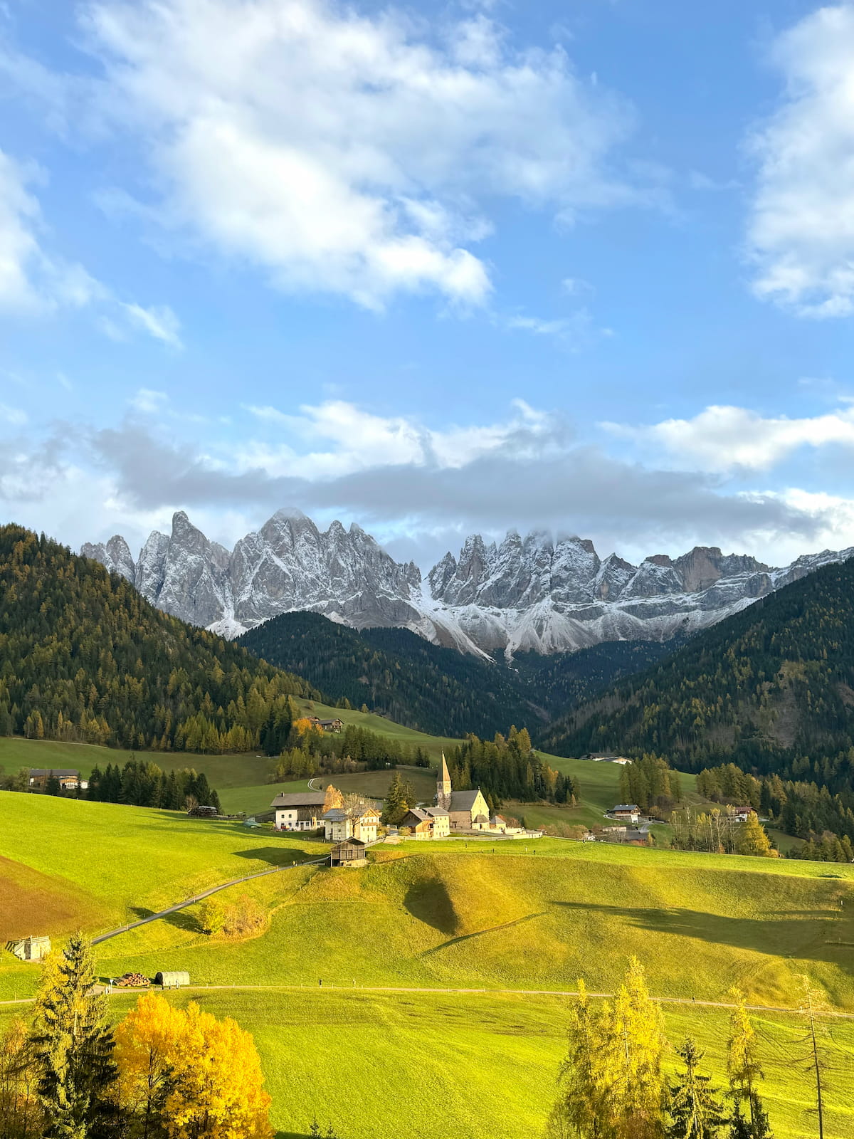 The historic Santa Maddalena church nestled in the rolling green hills of Val di Funes, with the dramatic, jagged Odle peaks of the Italian Dolomites rising into a clear blue sky.
